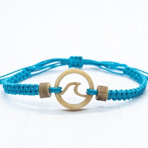 May include: A turquoise blue braided bracelet with a wooden circle charm featuring a carved wave design.