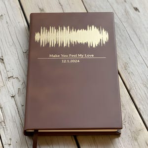 May include: A brown journal with gold foil accents. The cover features a sound wave design and the text "Make You Feel My Love" with the date 12.1.2024. A brown ribbon bookmark is visible.