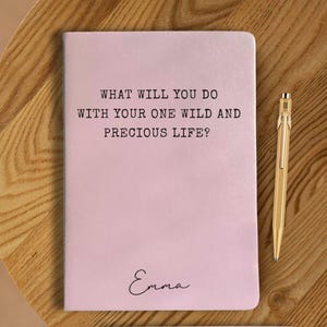 May include: A pale pink notebook with the text "WHAT WILL YOU DO WITH YOUR ONE WILD AND PRECIOUS LIFE?" and the name "Emma" in cursive. A gold pen lies beside the notebook on a wooden surface.