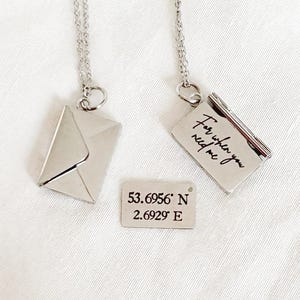 May include: Two silver necklaces with pendants shaped like an envelope and a book. The book pendant has the inscription "For when you need me." A small rectangular tag displays coordinates. The items are on a white surface.