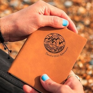 A brown, personalized journal being held in a hand. The cover is embossed with a unique travel design, along with your name and the phrase “wild adventures”.