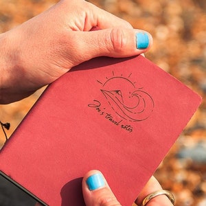 Puede incluir: Un diario de viaje de cuero rojo con un diseño de sol, ola y barco y el texto "This is travel notes".