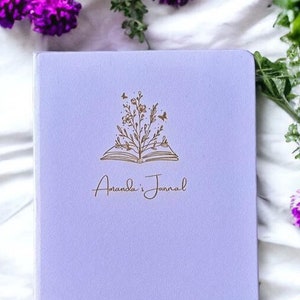 Op de afbeelding: Een lavendelkleurig notitieboek met een gouden bloemenontwerp en de tekst "Amanda's Journal" op de voorkant.