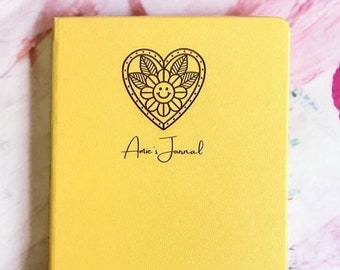 May include: A bright yellow notebook featuring a black line art graphic of a heart containing a flower with a smiley face. Below the heart, the text "Amie's Journal" is printed in a black cursive script font on the front cover.