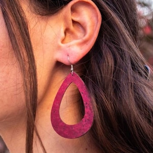 Puede incluir: Un par de pendientes en forma de lágrima hechos de corcho. Los pendientes son de color burdeos oscuro y tienen un diseño recortado en el centro. Los pendientes cuelgan de ganchos plateados.