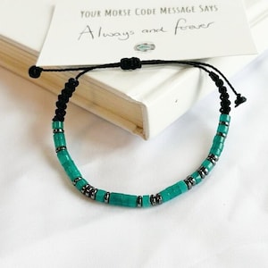 May include: A turquoise beaded bracelet with black cord and adjustable knot. The bracelet features cylindrical beads separated by small, dark metal spacers. A card with the text "Your Morse Code Message Says Always and forever" rests behind the bracelet.