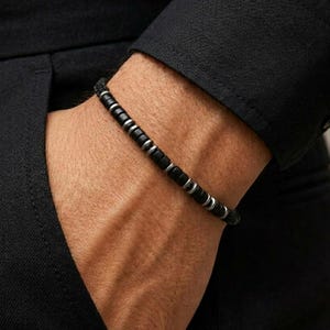 Op de afbeelding: Een kralenarmband met een herhalend patroon van kleine, matzwarte cilindrische kralen afgewisseld met gepolijste zilverkleurige metalen tussenstukjes, gedragen om de pols tegen een zwarte stoffen mouw.
