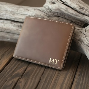 Puede incluir: Una cartera de cuero marrón con costuras blancas y las iniciales "MT" en dorado. La cartera es rectangular y está sobre una superficie de madera con madera flotante en el fondo. La cartera parece nueva.