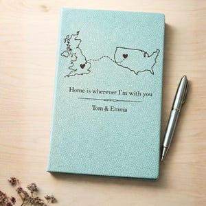 May include: A pale blue journal with a textured finish, showing a map of the UK and the USA linked by a dotted line. The cover reads "Home is wherever I'm with you" and the names "Tom & Emma". A silver pen lies next to the journal.
