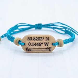 May include: A turquoise blue braided bracelet with wooden beads and a wooden tag that reads "50.8203&deg; N 0.1446&deg; W" with an arrow pointing to the right.