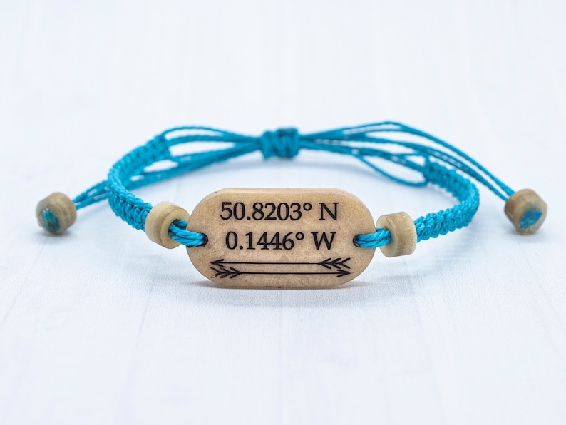 May include: A turquoise blue braided bracelet with wooden beads and a wooden tag that reads "50.8203&deg; N 0.1446&deg; W" with an arrow pointing to the right.