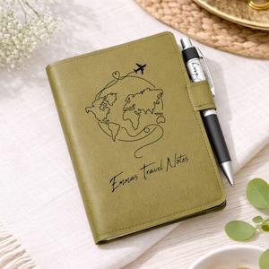 Puede incluir: Diario de viaje verde oliva con un diseño de mapa del mundo y avión. El diario tiene el texto "Emmas Travel Notes" e incluye un bolígrafo negro y plateado. El diario está sobre una tela blanca.
