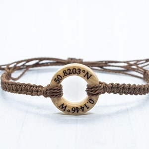May include: A brown braided bracelet with a wooden ring charm. The ring has the coordinates "50.8203&deg; N 0.1441&deg; O" engraved on it.