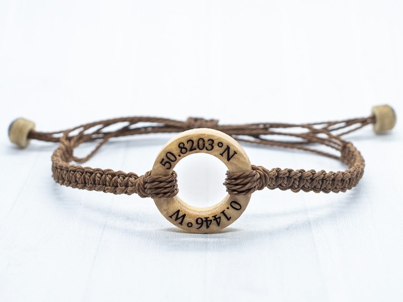 May include: A brown braided bracelet with a wooden ring charm. The ring has the coordinates "50.8203&deg; N 0.1441&deg; O" engraved on it.