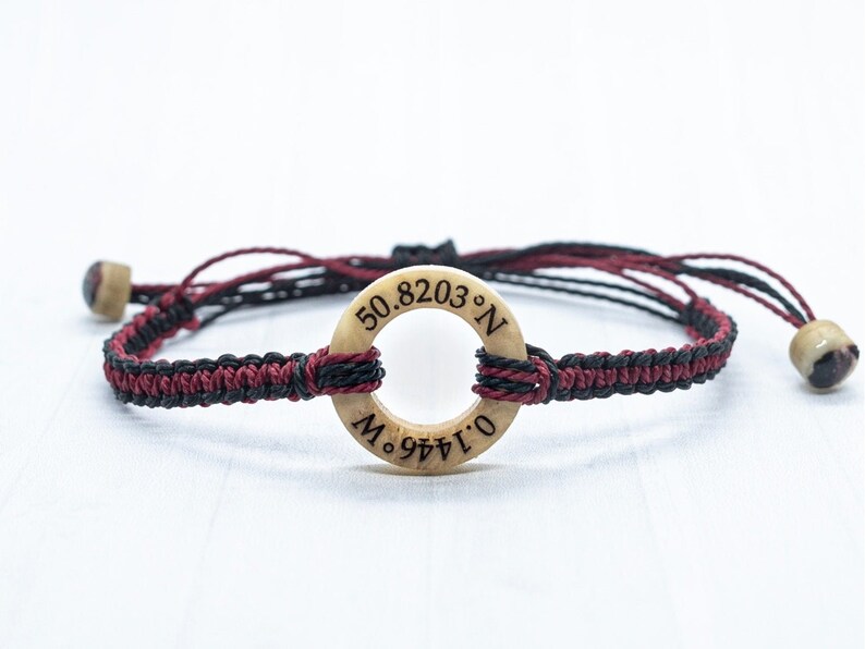 May include: A black and burgundy braided bracelet with a wooden circle charm. The charm has the coordinates 50.8203&deg; N and 0.1441&deg; E engraved on it.