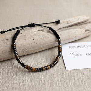 Può includere: Un braccialetto di perline marroni e argentate, con un cordino nero regolabile. Il braccialetto è esposto su un pezzo di legno trasportato dalla corrente, accanto a un biglietto bianco con la scritta "You're my".