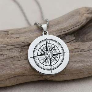 May include: A silver-colored pendant necklace featuring a compass rose design. The pendant is round with a detailed compass rose graphic in black. The necklace is displayed on a piece of driftwood.