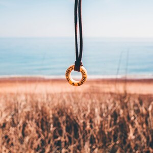 May include: A brown leather cord necklace with a round wooden pendant. The pendant has the coordinates 43.8008&deg; N 101.6916&deg; E engraved on it. The necklace is hanging in front of a blurred background of a beach and ocean.