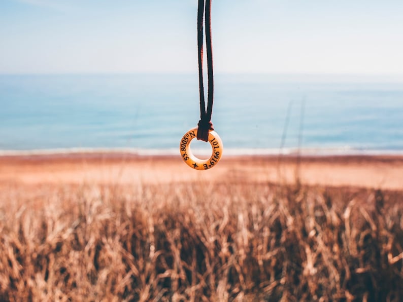 May include: A brown leather cord necklace with a round wooden pendant. The pendant has the coordinates 43.8008&deg; N 101.6916&deg; E engraved on it. The necklace is hanging in front of a blurred background of a beach and ocean.