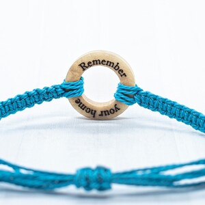 May include: A turquoise blue friendship bracelet with a wooden circle charm that says "Remember Your Home".