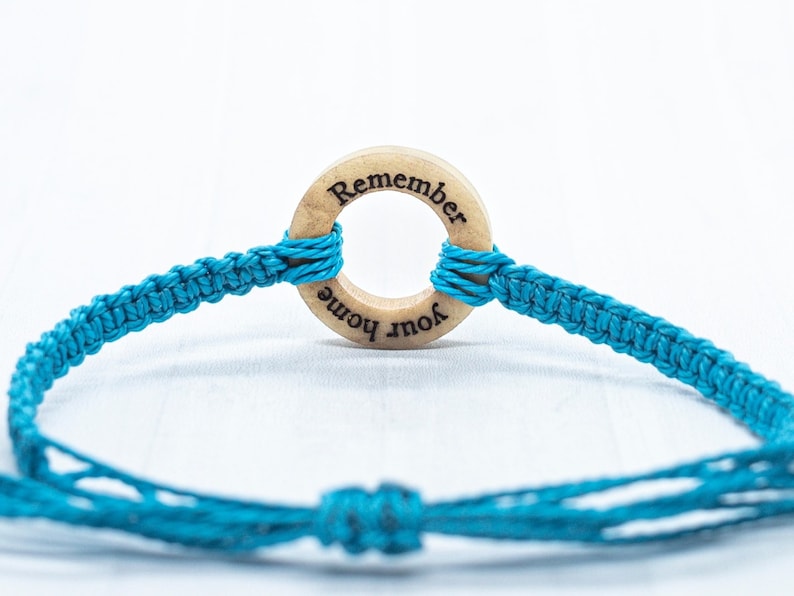 May include: A turquoise blue friendship bracelet with a wooden circle charm that says "Remember Your Home".