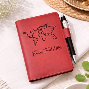 May include: A red leather travel journal with a world map design and the text "Emmas Travel Notes". A black pen is attached to the side. The journal is a perfect gift for travelers.