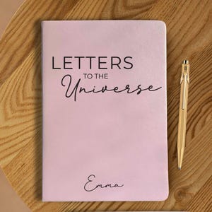 May include: A light pink notebook with the text "LETTERS TO THE Universe" and the name "Emma" in black script. A gold pen rests on a wooden surface next to the notebook. The notebook is a journal.