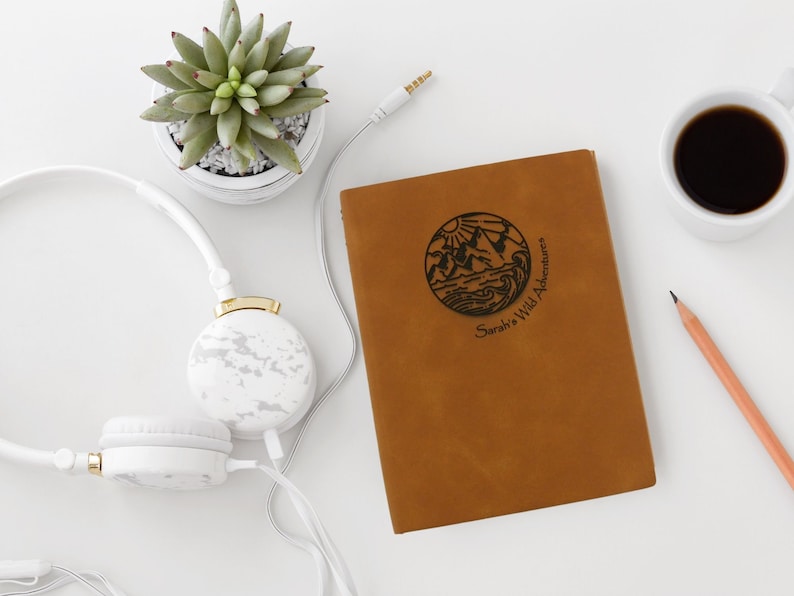 A brown, personalized journal stylishly arranged on a table. The cover is engraved with a unique travel notebook design, along with your name and the phrase “wild adventures”.