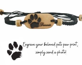 May include: Black braided bracelet with a beige oval charm featuring a paw print and the name "Dennis" engraved on it.  The charm is attached to the bracelet with two beige beads.