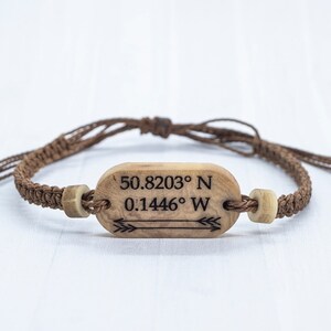 May include: A brown cord bracelet with a wooden tag that has the coordinates "50.8203&deg; N 0.1446&deg; W" and an arrow engraved on it.