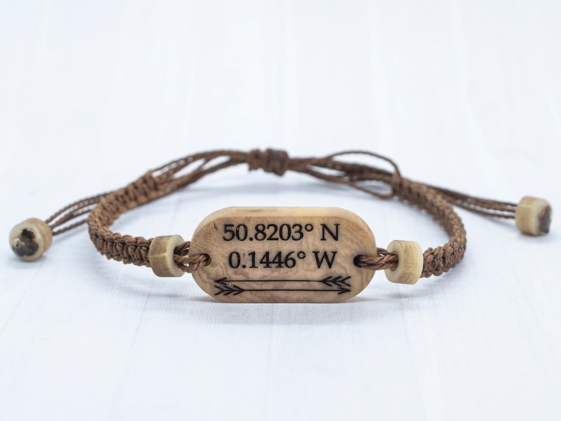 May include: A brown cord bracelet with a wooden tag that has the coordinates "50.8203&deg; N 0.1446&deg; W" and an arrow engraved on it.