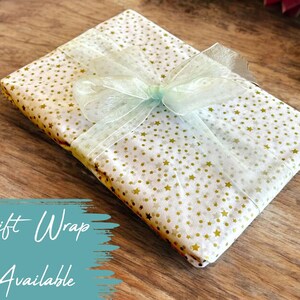 May include: A white gift box with a gold star pattern is wrapped with a light green ribbon. The box is sitting on a wooden surface.