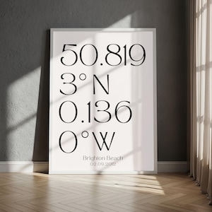 May include: A framed white print with black text featuring coordinates and the words "Brighton Beach 02.09.2012". The coordinates are 50.819, 3° N, 0.136, and 0° W. The print is displayed on a wooden floor with a gray wall and a curtain.