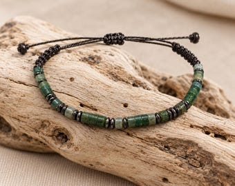 Puede incluir: Una pulsera que presenta una alternancia de cuentas cilíndricas de piedra verde y pequeñas cuentas espaciadoras metálicas de color gris oscuro. La pieza está ensartada en un cordón negro fino con cierre de nudo corredizo.