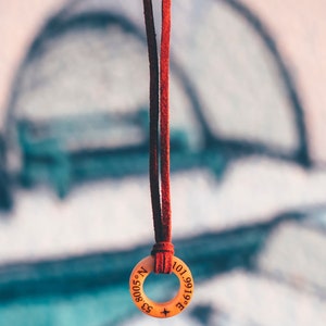 May include: A red leather cord necklace with a wooden pendant. The pendant is round and has the coordinates "53.8005&deg; N 101.9919&deg; E" engraved on it.