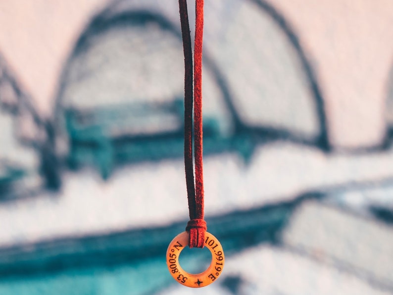 May include: A red leather cord necklace with a wooden pendant. The pendant is round and has the coordinates "53.8005&deg; N 101.9919&deg; E" engraved on it.