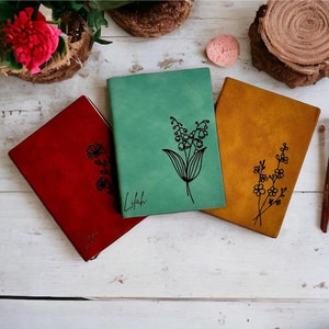 Puede incluir: Tres diarios encuadernados en cuero con diseños florales. Los diarios son rojo, verde y marrón. El diario rojo tiene un diseño floral con la palabra "You" inscrita en él. El diario verde tiene un diseño floral con la palabra "Lylah" inscrita en él. El diario marrón tiene un diseño floral.