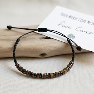 May include: A black cord bracelet with brown and silver beads. A white card with the text "Your Morse Code Message Says Fuck Cancer" rests beside the bracelet on a wooden surface. The bracelet is adjustable.