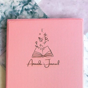 Peut inclure: Un journal rose avec une illustration en noir et blanc d'un livre ouvert avec des fleurs et des étoiles. Le texte "Amanda's Journal" est écrit en cursive sous l'illustration.