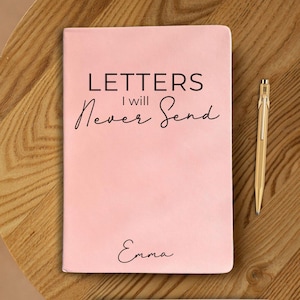 May include: A pink notebook with the text "LETTERS I will Never Send" in black script. The name "Emma" is written at the bottom. A gold pen rests beside the notebook on a wooden surface.