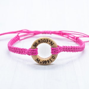 May include: Pink braided bracelet with a wooden circle charm engraved with the coordinates "50.8203&deg; N, 0.1444&deg; E".