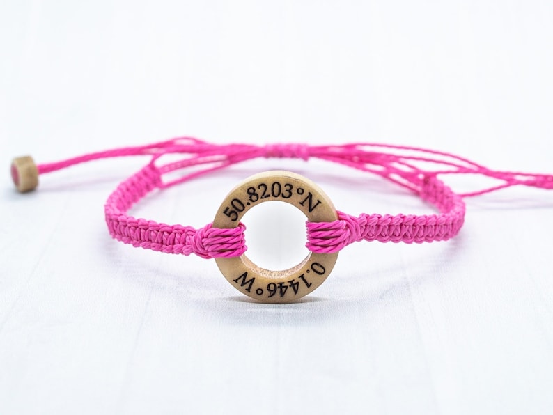 May include: Pink braided bracelet with a wooden circle charm engraved with the coordinates "50.8203&deg; N, 0.1444&deg; E".