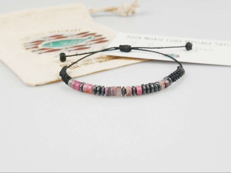 Pink jade and Hematite bracelet with customised Morse code message, photographed with branded hemp gift bag and message card blurred in the background.