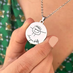 Op de afbeelding: Een zilveren ketting met een ronde hanger met een kinderlijke tekening van een stokfiguur met de woorden "love you daddy" erop.