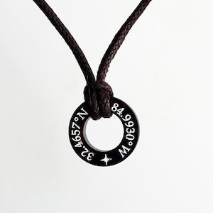 May include: A black cord necklace with a black metal ring pendant. The ring has white engraved text that reads "32.4657&deg; N. 84.9930&deg; W."