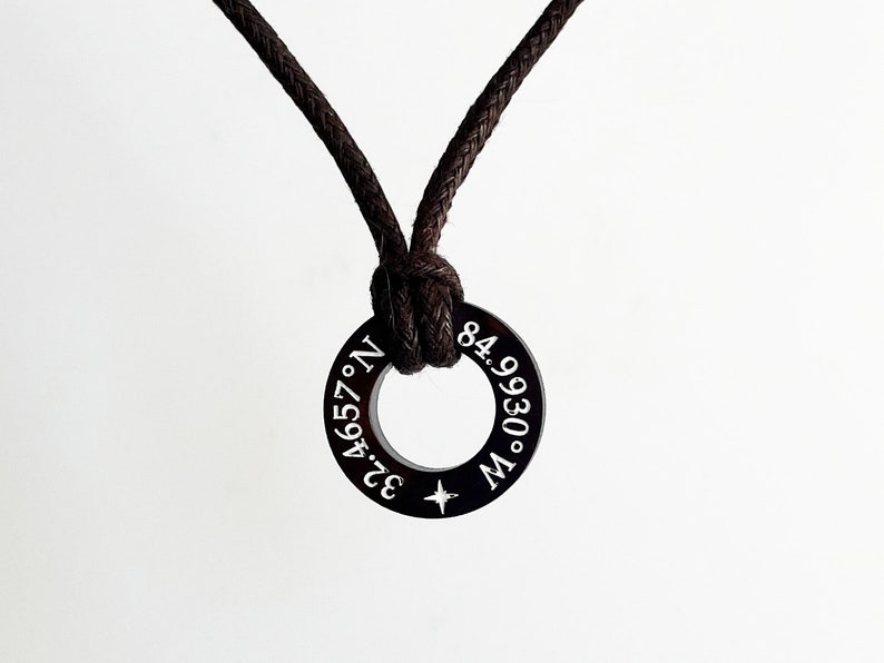 May include: A black cord necklace with a black metal ring pendant. The ring has white engraved text that reads "32.4657&deg; N. 84.9930&deg; W."