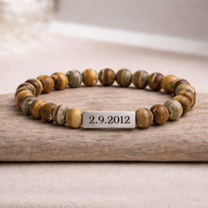 May include: A beaded bracelet with brown, tan, and grey beads. A rectangular silver plate is engraved with the date "2.9.2012". The beads are strung together, forming a circular bracelet. The bracelet is displayed on a wooden surface.