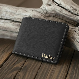 Puede incluir: Cartera de cuero negro con costuras blancas y la palabra "Daddy" en letras doradas. La cartera es rectangular y está sobre una superficie de madera.