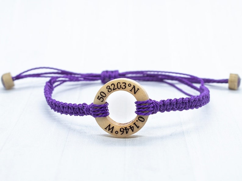 May include: Purple braided bracelet with a wooden circle charm engraved with the coordinates "50.8203&deg; N 0.1446&deg; E".