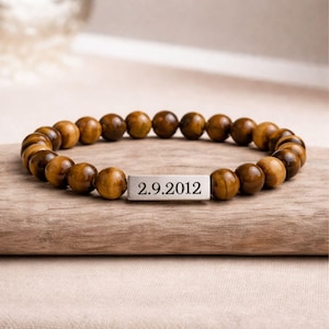 May include: A bracelet featuring round, tiger-eye beads. A silver rectangular charm displays the date "2.9.2012". The bracelet is presented on a wooden surface.